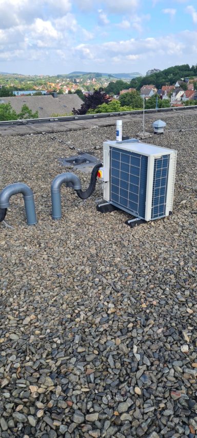 Klimaanlage auf einem Flachdach, umgeben von Rohren und Kies.