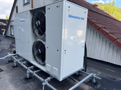 Klimaanlage mit zwei Lüftern auf einem Dach mit Haus im Hintergrund.